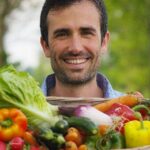 Mann mit Korb voller frischem Gemüse wie Paprika, Auberginen, Radieschen, Karotten und Salatblättern draußen.