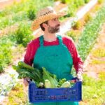 Mann mit Strohhut und grünem Overall auf Feld trägt Kiste mit frischem Gemüse.
