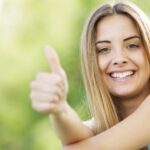 Junge Frau mit langen blonden Haaren zeigt Daumen hoch vor unscharfem grünem Hintergrund.