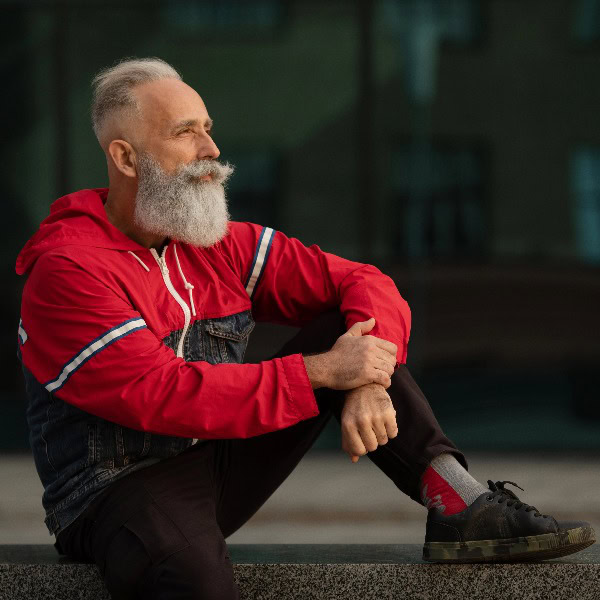 Älterer Mann mit weißem Vollbart sitzt auf einer Mauer. Er trägt eine rote Jacke und dunkelbraune Jeans. Er schaut zufrieden zur Seite.