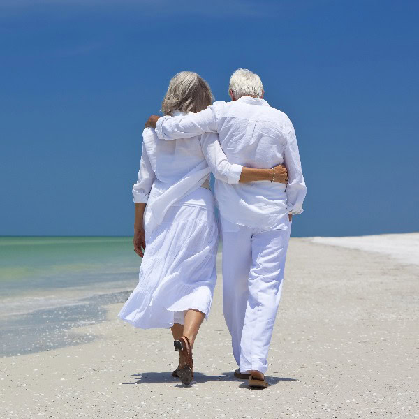 Ein älteres Paar in weißer Kleidung läuft am Strand entlang. Wir sehen sie von hinten.