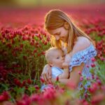 Junge Frau sitzt mit ihrem Baby in einem Feld voller Rosen. Das Baby bekommt die Brust.
