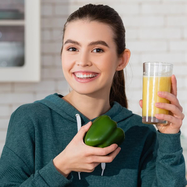 Junge lächelnde Frau in blaugrünem Pullover mit Paprika und leckerem Smoothie in den Händen.