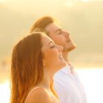 Mann und Frau stehen mit geschlossenen Augen, Kopf zum Himmel erhoben, genießen die Sonne und atmen frei ein.
