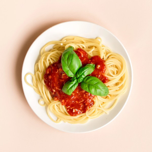 Teller mit Spaghettinudeln und Tomatensauce und Basilikumblättern auf beigem Untergrund.